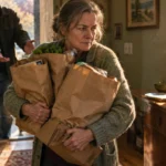 A woman carrying heavy paper grocery bags indoors looks away as a man reaches out to offer help.