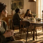 A woman sitting on a sofa holding a mug observes a group of people conversing around a dining table.