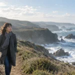 A woman in a grey coat and scarf walks along a sunny coastal cliff path overlooking crashing ocean waves.