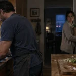 Man washes dishes at a kitchen sink while a woman carrying groceries stands near an open refrigerator looking toward him.